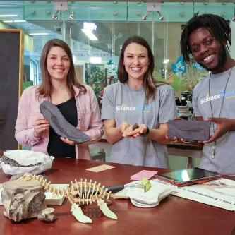 Two light-skinned women and one dark-skinned man holding fossils while standing behind a table with fossils on it, including what looks like a model of a skeleton of a long-necked creature with flippers. A large fossil of a similar creature embedded in stone stands upright to the left of the people.