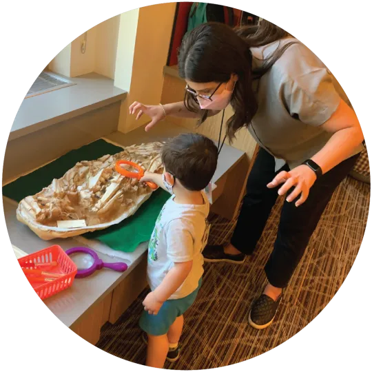 A boy holding a magnifying glass over a cast of a fossil bed, while a woman stands over him making scary monster arms