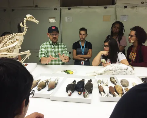 Six teenagers standing around a table that more than a dozen taxidermic birds on it, a taxidermic snowy owl, and a skeleton of a large wading bird. A man in a black baseball cap is gesturing as he speaks to the group.