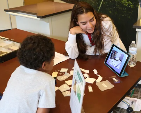 A teen girl sitting at a table, making a presentation to a younger child. An iPad displaying a photo is propped on the table and there are objects and scraps of paper on the table.