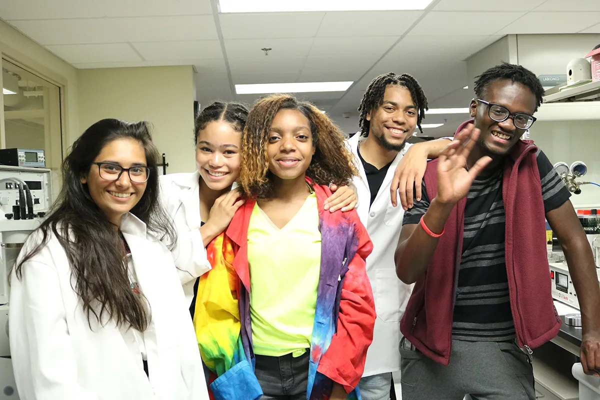 Five teenagers — three girls and two boys — standing together, smiling, in a science lab. Three of the teens are wearing white lab coats. One boy is waving.