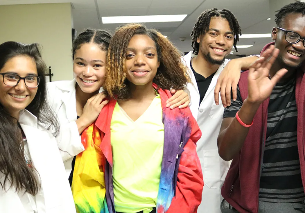 Five teenagers -- three girls and two boys -- standing together, smiling, in a science lab. Three of the teens are wearing white lab coats. One boy is waving.