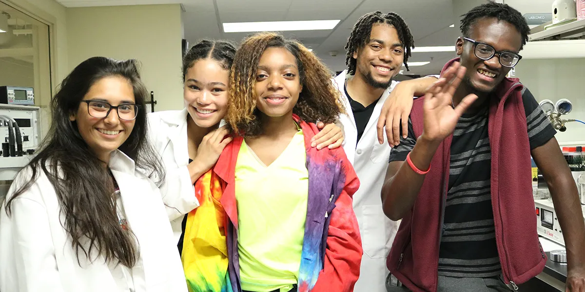 Five teenagers -- three girls and two boys -- standing together, smiling, in a science lab. Three of the teens are wearing white lab coats. One boy is waving.