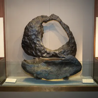 Two very large pieces of blackish-brown rock in a museum display case. The top one is gumdrop-shaped and has a large hole in it and its surface looks smooth, but has many indentations. The bottom one is just as wide as the top piece, but only one-third as tall with no holes. It also has a smooth surface with indentations. 