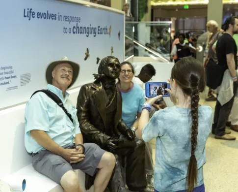 A girl takes a photo of two adults sitting by a bronze of Charles Darwin