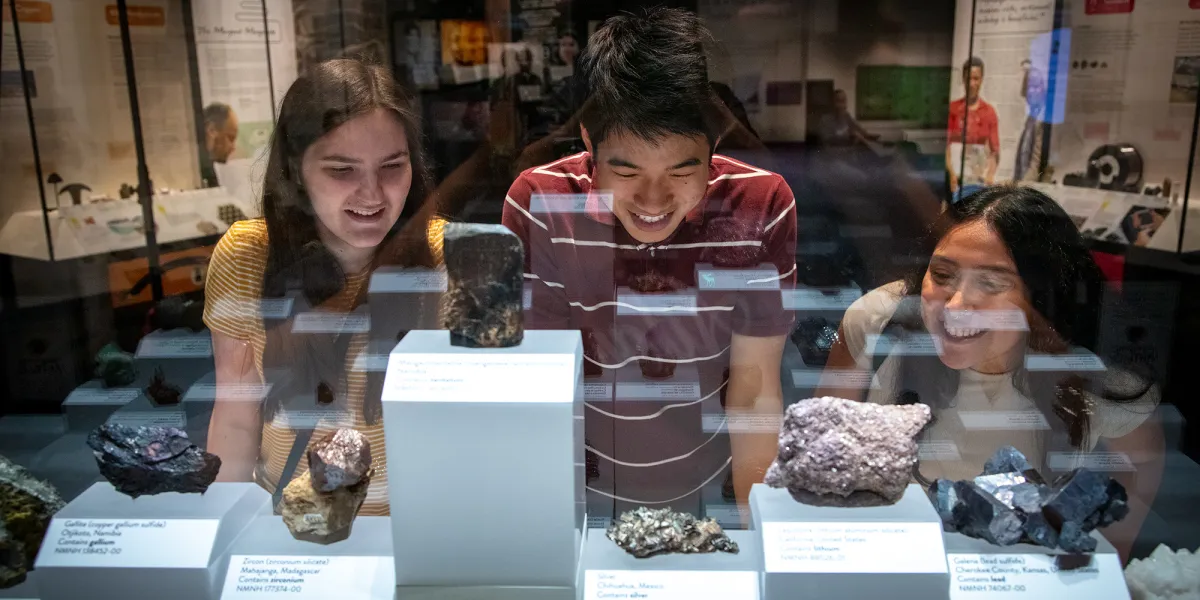 Three teenagers look into an exhibit case of minerals, as seen from through the case on the other side. There are eight minerals in the image, of various sizes and shapes and ranging in color from white to black.