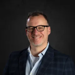 A light-skinned man with dark hair, black-rimmed glasses, and a slight beard
